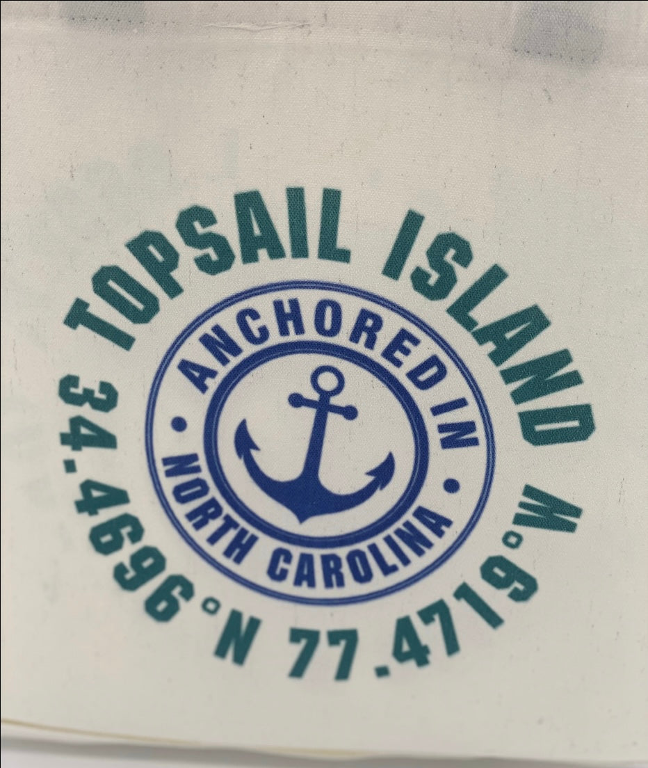 White tote bag with black handles, featuring a circular design with text reading 'TOPSAIL ISLAND,' 'ANCHORED IN,' and 'NORTH CAROLINA,' along with an anchor symbol and geographic coordinates.