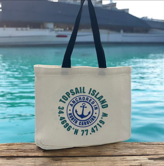 White tote bag with black handles, featuring a circular design with text reading 'TOPSAIL ISLAND,' 'ANCHORED IN,' and 'NORTH CAROLINA,' along with an anchor symbol and geographic coordinates.