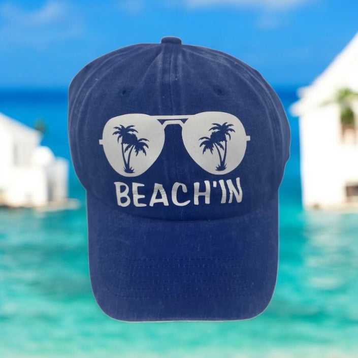 Navy blue cap with white 'BEACH'IN', sunglass, and palm tree design against a beach background