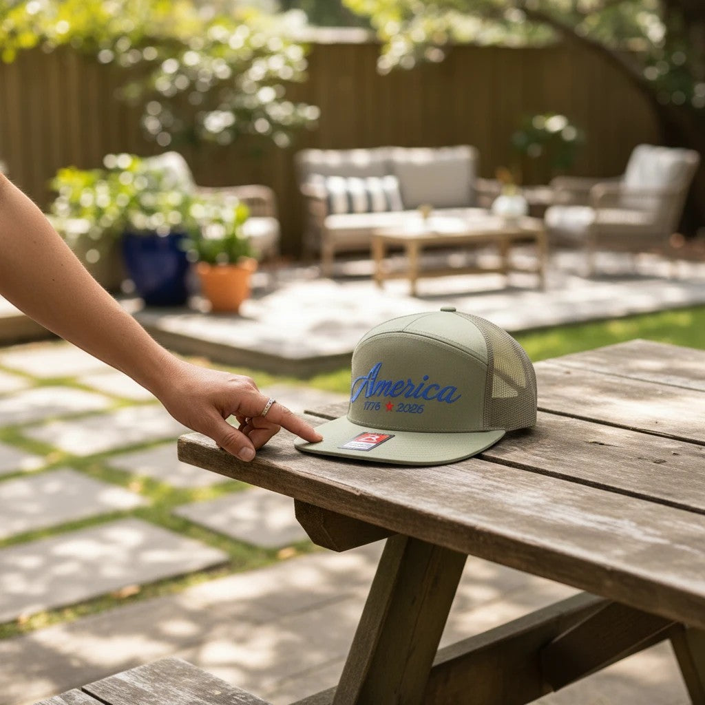 Seaside Custom Apparel & Gifts olive green Richardson trucker hat with America 1776-2026 embroidery on a wooden table