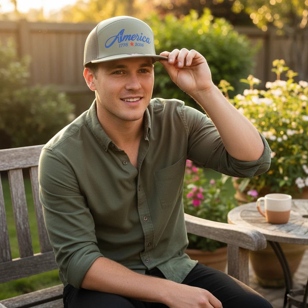 Man wearing a Seaside Custom Apparel & Gifts olive green trucker hat with America embroidery sitting on a bench