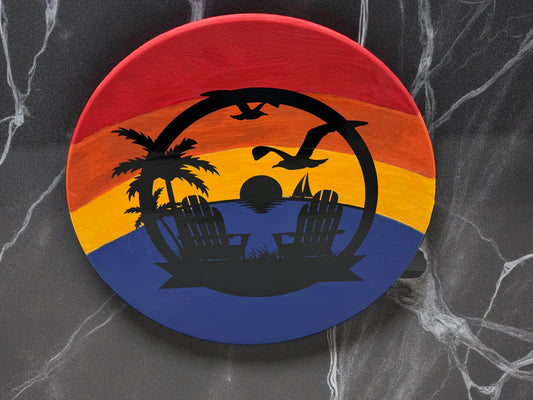 Round plaque with a vibrant sunset gradient and black beach silhouette featuring palm trees, lounge chairs, birds, and a setting sun, with personalized ribbon below the chairs.
