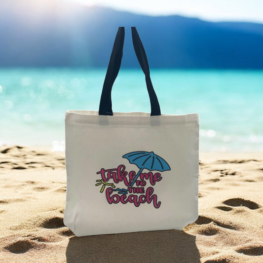 Tote bag with 'take me to the beach' text and umbrella graphic on sandy beach with ocean background